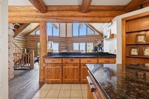 A log cabin interior with a kitchen area featuring wooden cabinets, a granite countertop, and a view through large windows.