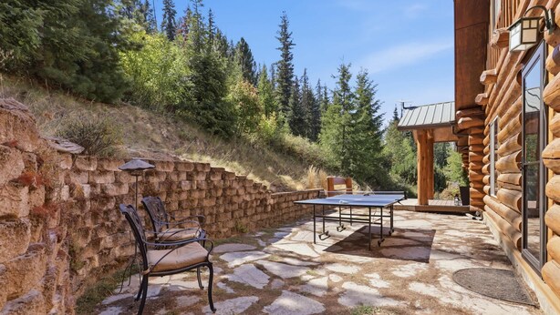 Un patio de piedra con mesa de ping pong y sillas, un muro de ladrillo y una cabaña de madera con techo inclinado.