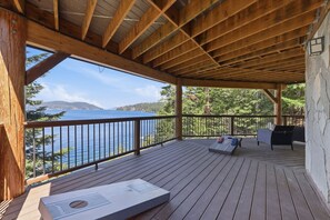 Terraza de madera con vistas a un lago y montañas, una parrilla y mobiliario de exterior.