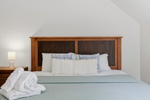 Un dormitorio con cabecera de madera, cama con almohadas blancas y estampadas, lámpara de noche y una toalla blanca enrollada en el suelo.