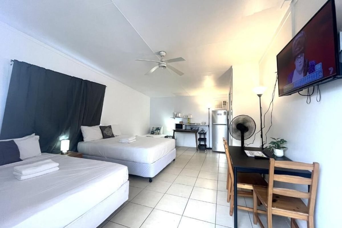 Habitación de hotel con dos camas, un escritorio, una silla, un televisor y una kitchenette al fondo.
