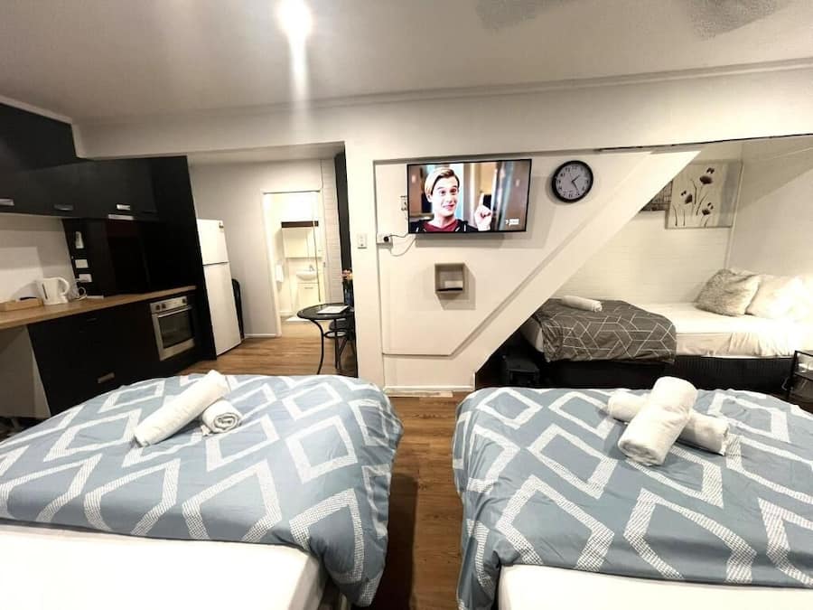 A compact hotel room with two beds, a kitchenette, a TV, and a clock on the wall.