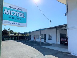 Un motel con un letrero que indica 'Aqua Blue Motel' y un número de teléfono. El edificio tiene techo plano y una zona de estacionamiento techada.