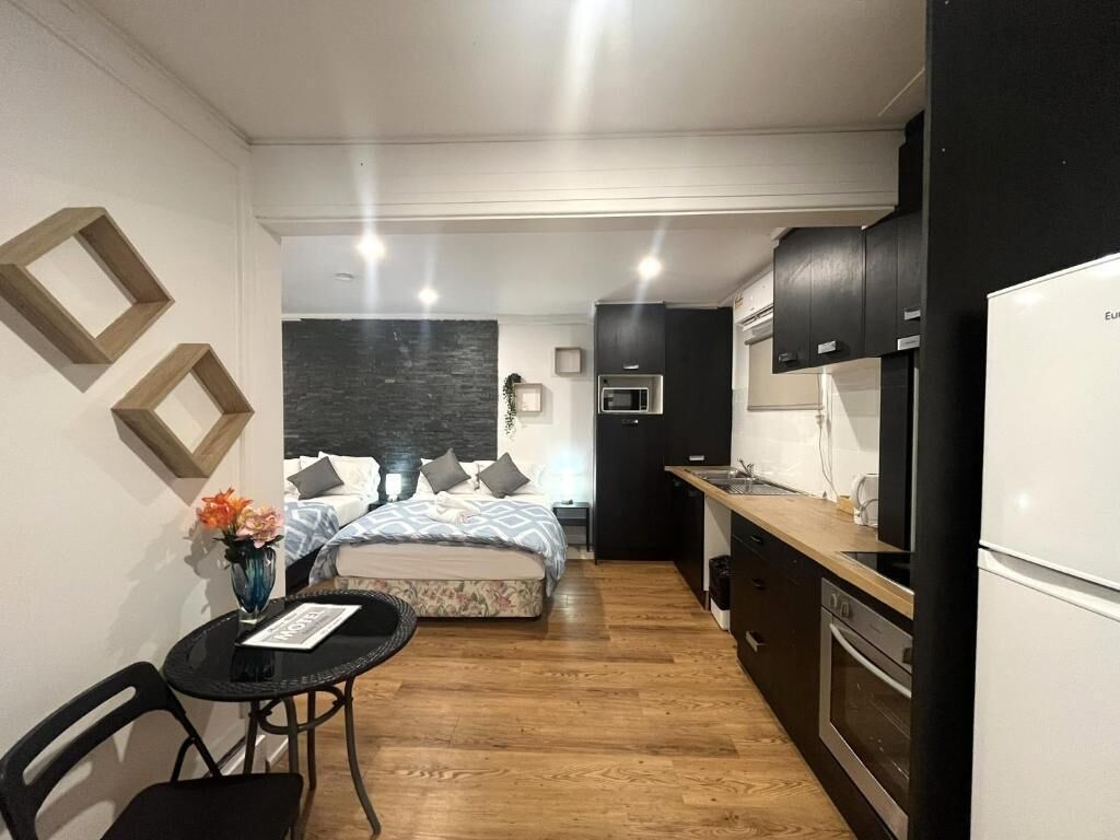 A compact hotel room with a kitchenette, a bed with patterned bedding, a small table with a vase, and a chair.