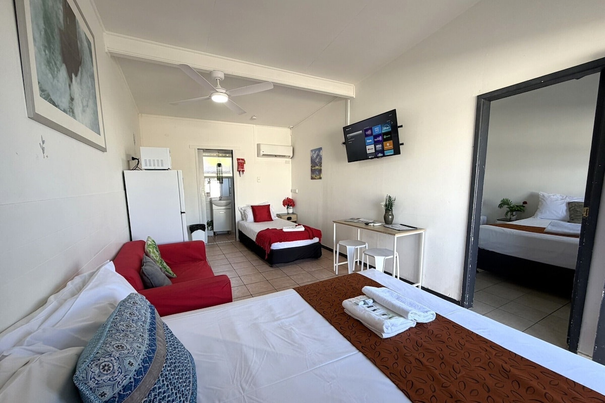 Habitación de hotel con cama, escritorio, silla, televisión y baño visible al fondo.