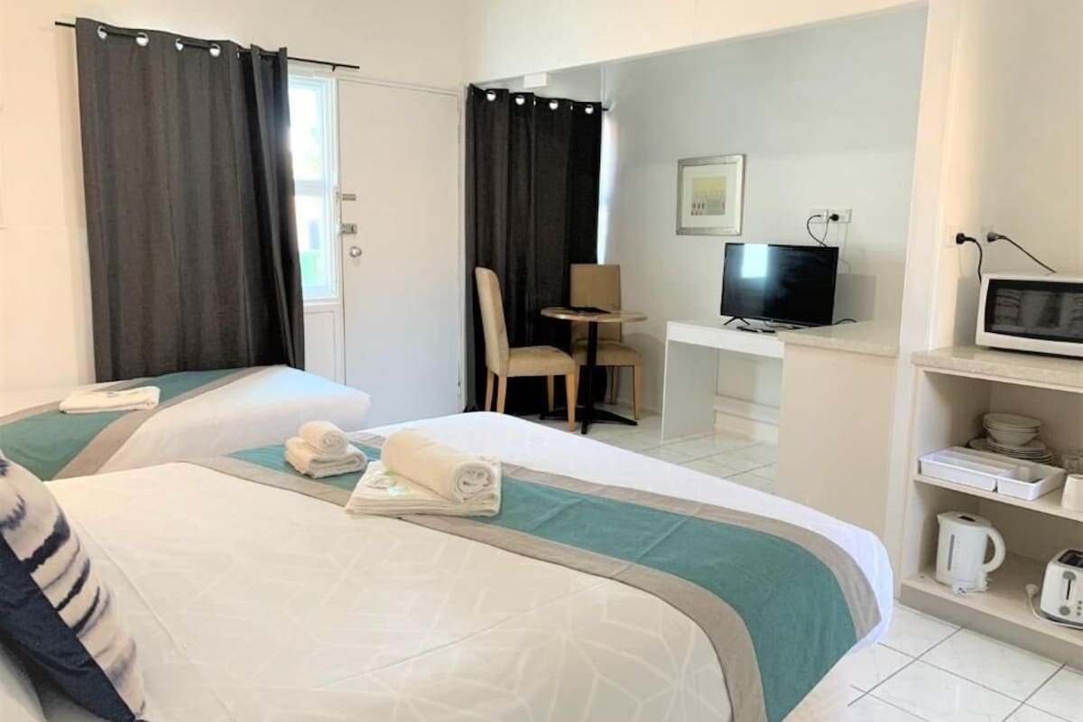 Habitación de hotel con dos camas, un escritorio con televisor, una silla y un estante con utensilios de cocina.