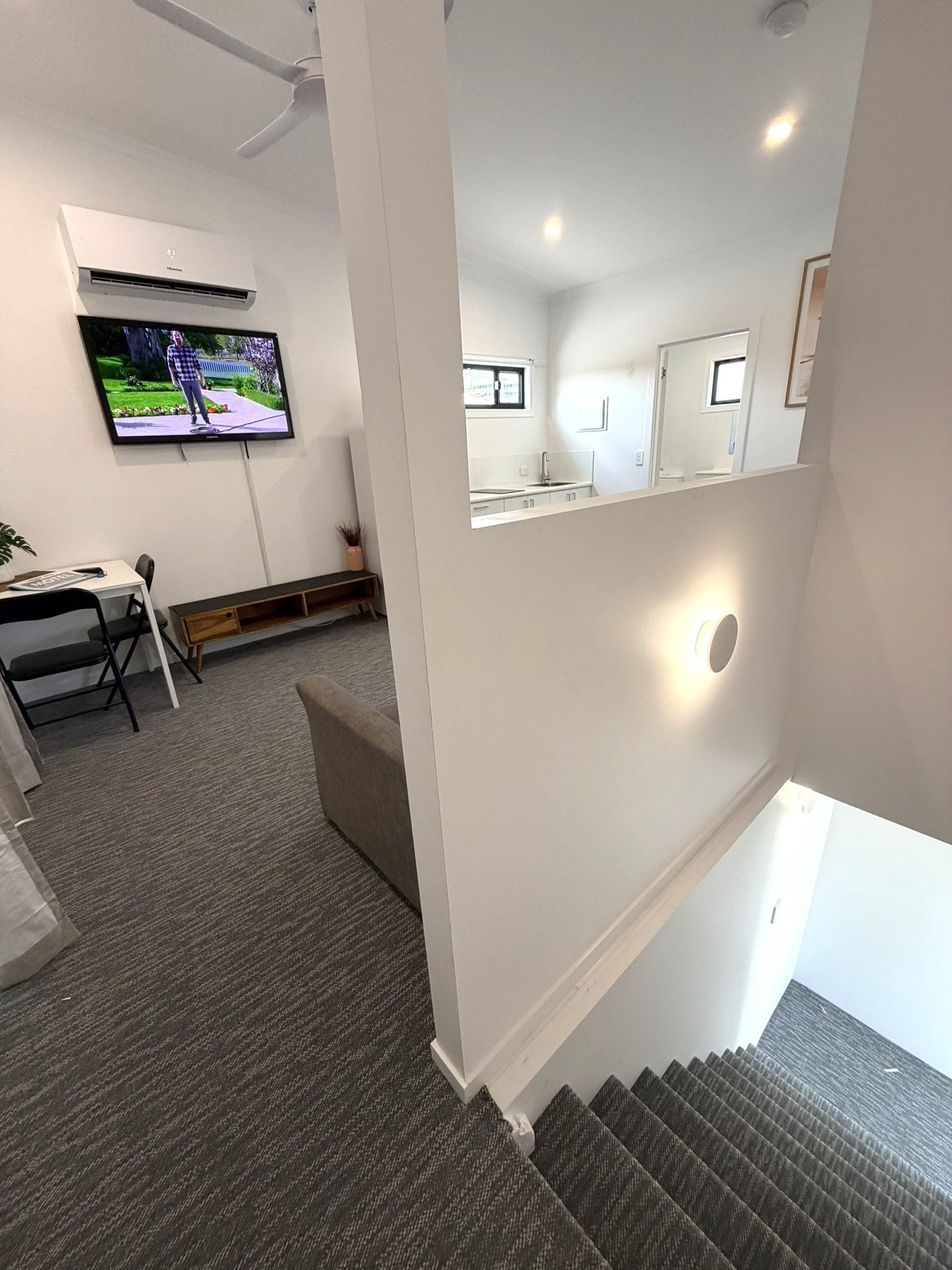 A modern hotel room with a flat-screen TV mounted on the wall, a desk with a chair, a small seating area, and a staircase leading to an upper level.