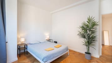 A bedroom with a bed, two bedside tables, a potted plant, and a door leading to another room.