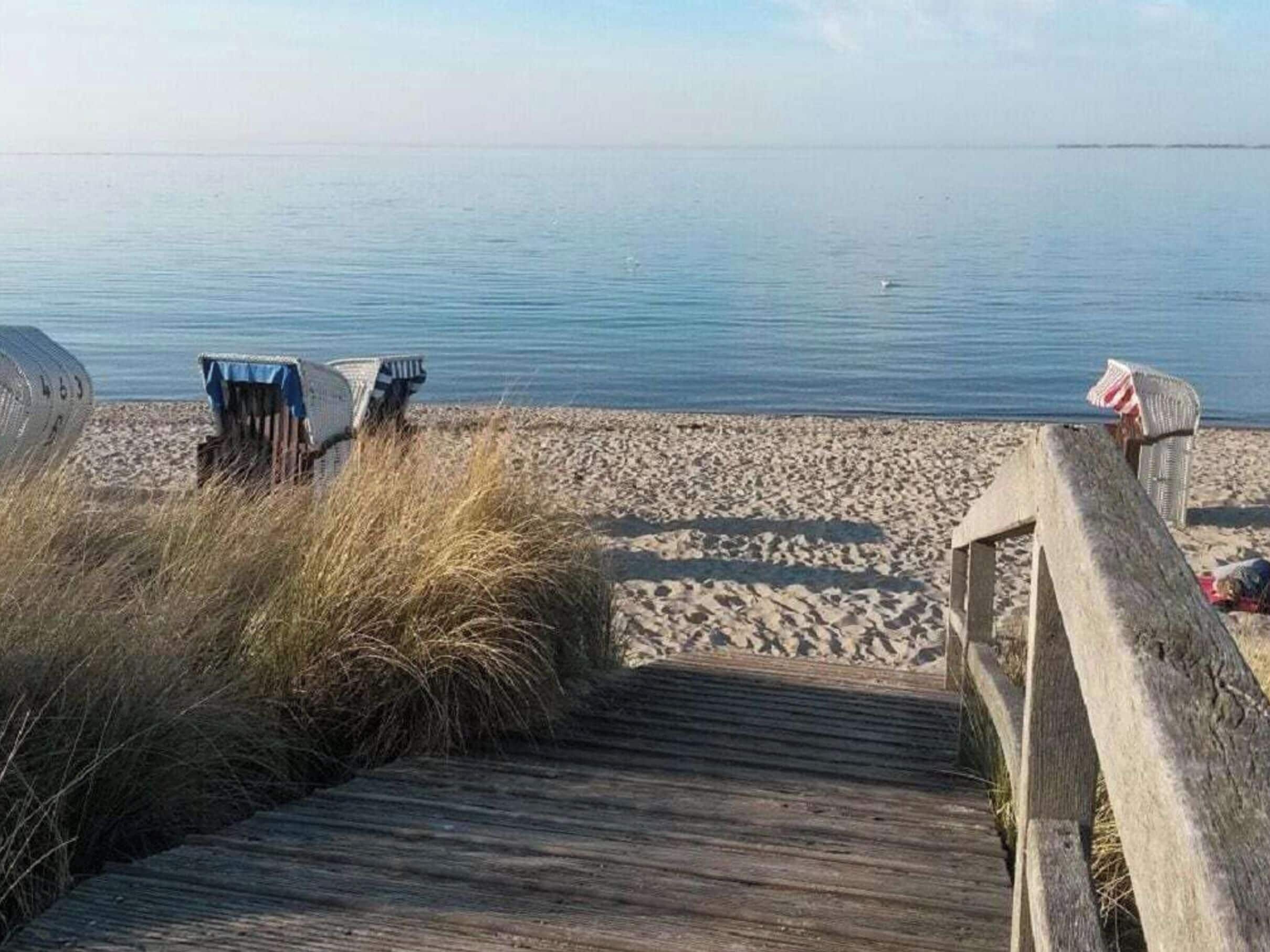Ein hölzerner Steg führt zu einem Sandstrand mit Liegestühlen und einer kleinen Konstruktion.