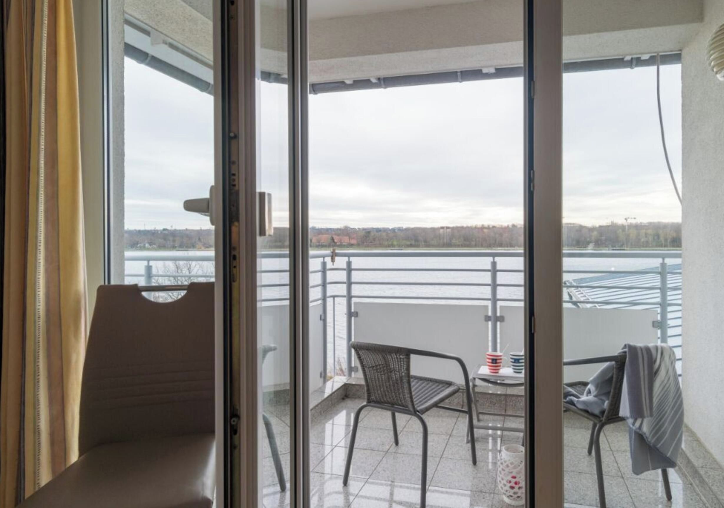 A balcony with a view of a body of water and a distant shoreline, furnished with a chair and a small table.