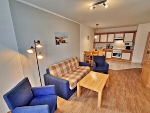 Ein Wohnzimmer mit Sofa, zwei Sesseln und einem Couchtisch. Es gibt eine Küchenzeile mit Schränken und einem Esstisch.