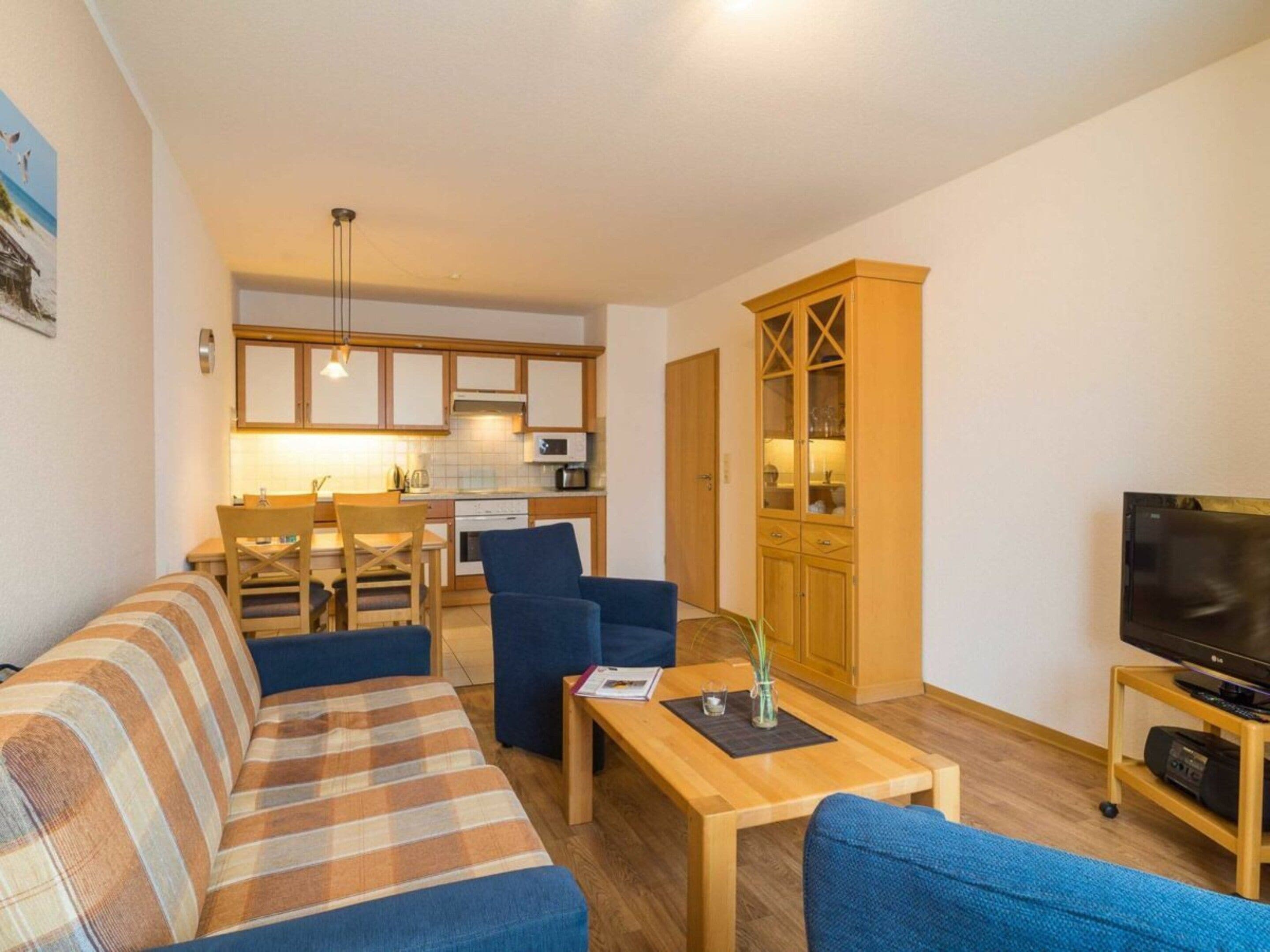 Sala de estar com sofá, cadeiras, mesa de centro, televisão e cozinha ao fundo.
