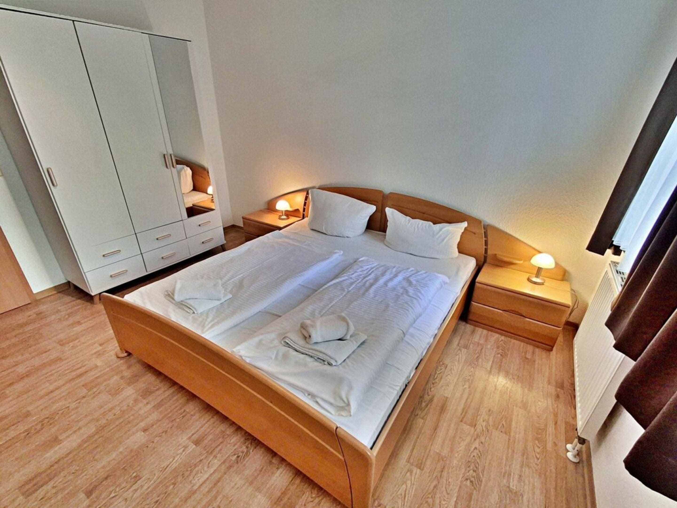 Une chambre d’hôtel avec un grand lit, une tête de lit en bois, deux tables de chevet avec des lampes, une armoire et un radiateur.
