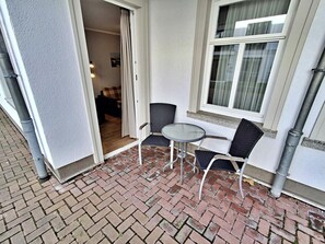 Un patio esterno con pavimento in mattoni, due sedie, un tavolino rotondo e una finestra con tende.