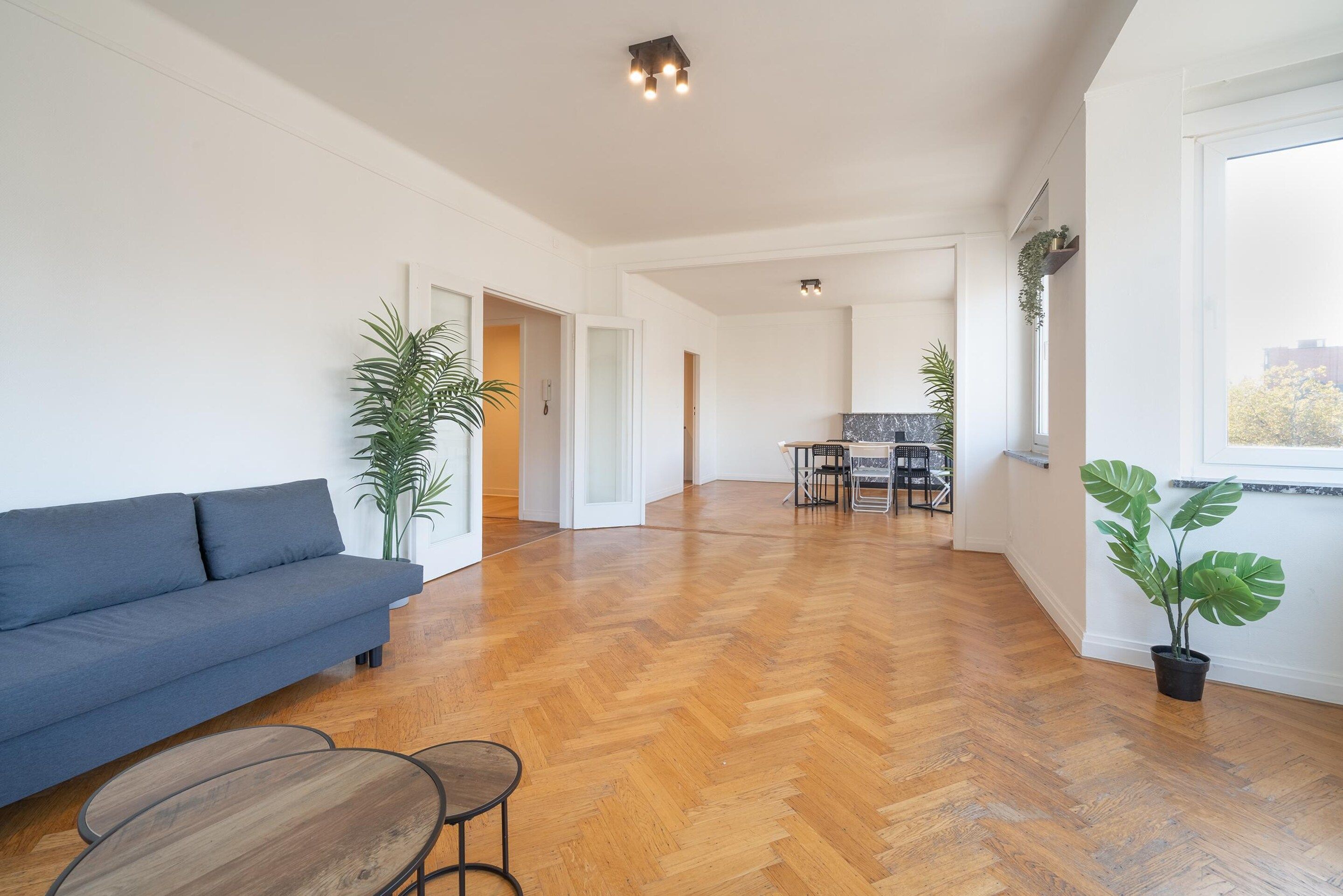 A spacious living area with a sofa, a dining table, and chairs, a kitchen in the background, and a large window with a view.