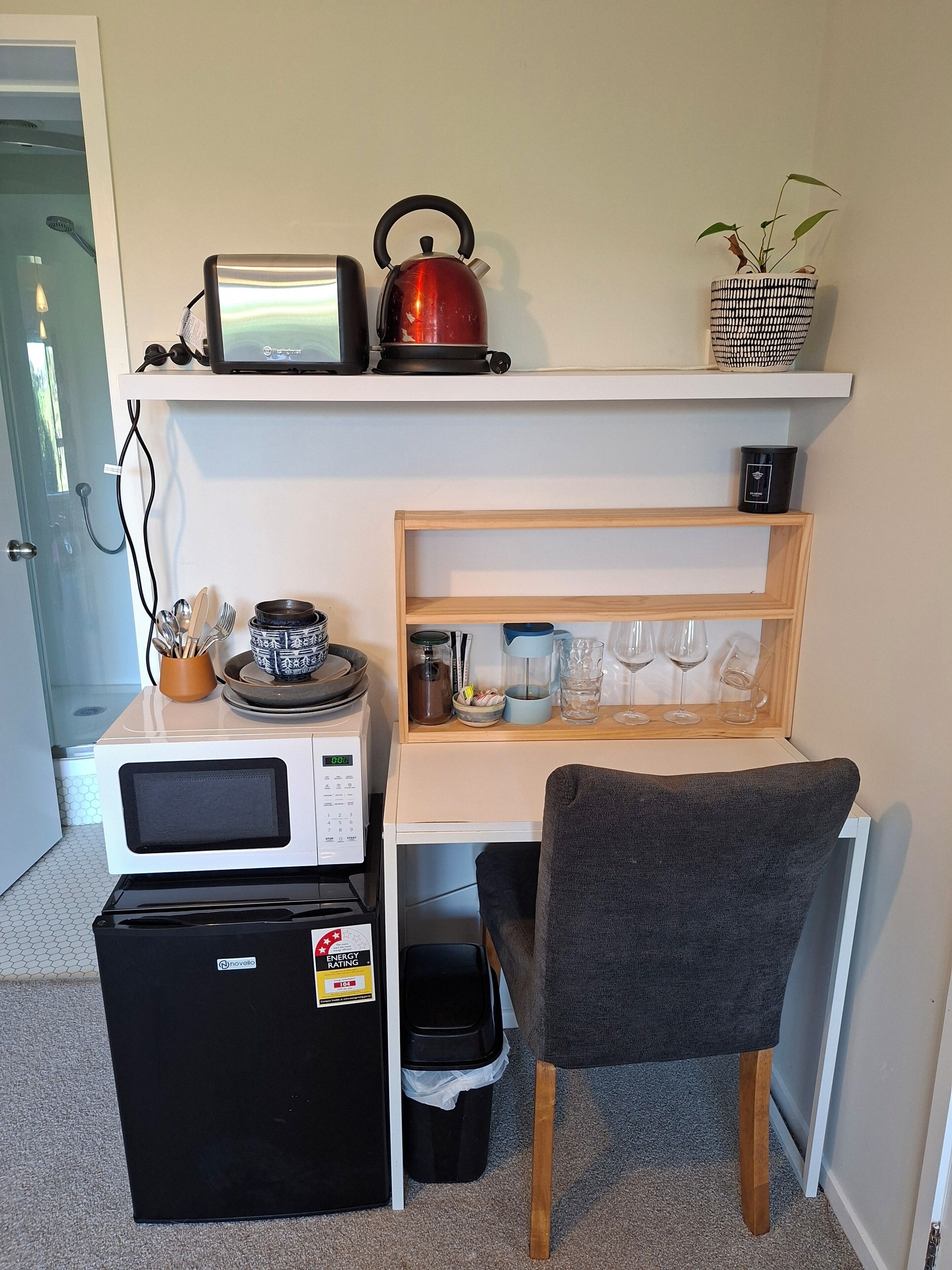 A compact kitchenette with a microwave, kettle, and small refrigerator. A chair is positioned in front of the counter, and a shelf above holds a kettle, a basket, and a small plant.