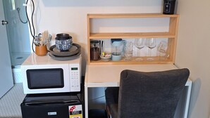 Une petite cuisine équipée d’un four à micro-ondes, d’une bouilloire et d’un petit réfrigérateur. Une chaise est placée devant le comptoir, et une étagère au-dessus contient une bouilloire, un panier et une petite plante.