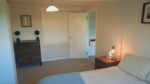 Quarto com cama, cômoda, mesa de cabeceira com abajur, porta e luminária no teto.