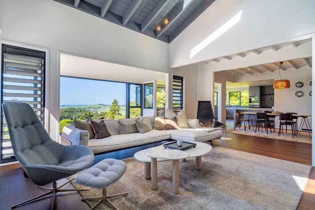 A spacious living area with a large sofa, a coffee table, and a chair. There is a view of the outdoors through large windows.
