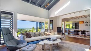 Un amplio espacio habitable con un sofá grande, una mesa de centro y una silla. Se aprecia vista al exterior a través de amplias ventanas.