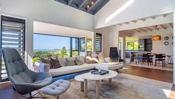 A spacious living area with a large sofa, a coffee table, and a chair. There is a view of the outdoors through large windows.