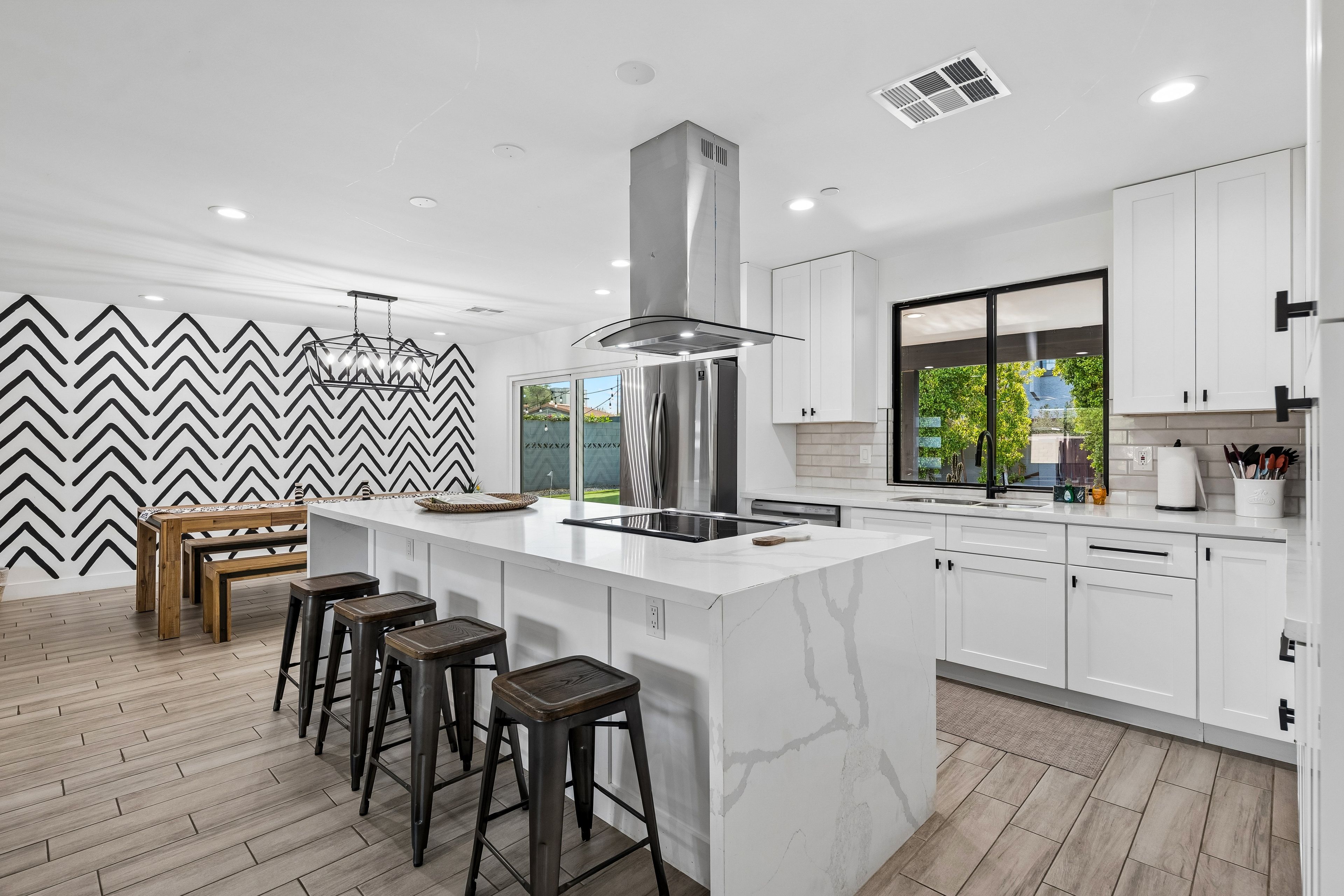A modern kitchen with a central island, bar stools, and a view through a window.