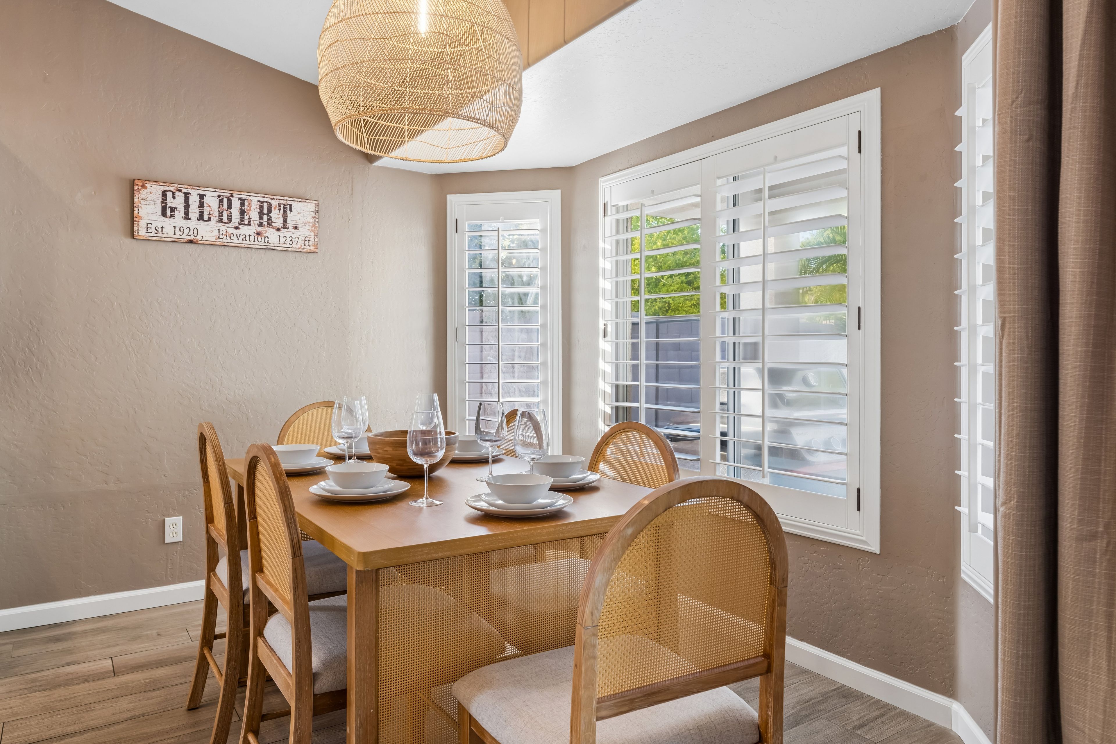 Een eethoek met een tafel gedekt voor vier personen, omringd door rieten stoelen, een hanglamp en een raam met luiken.