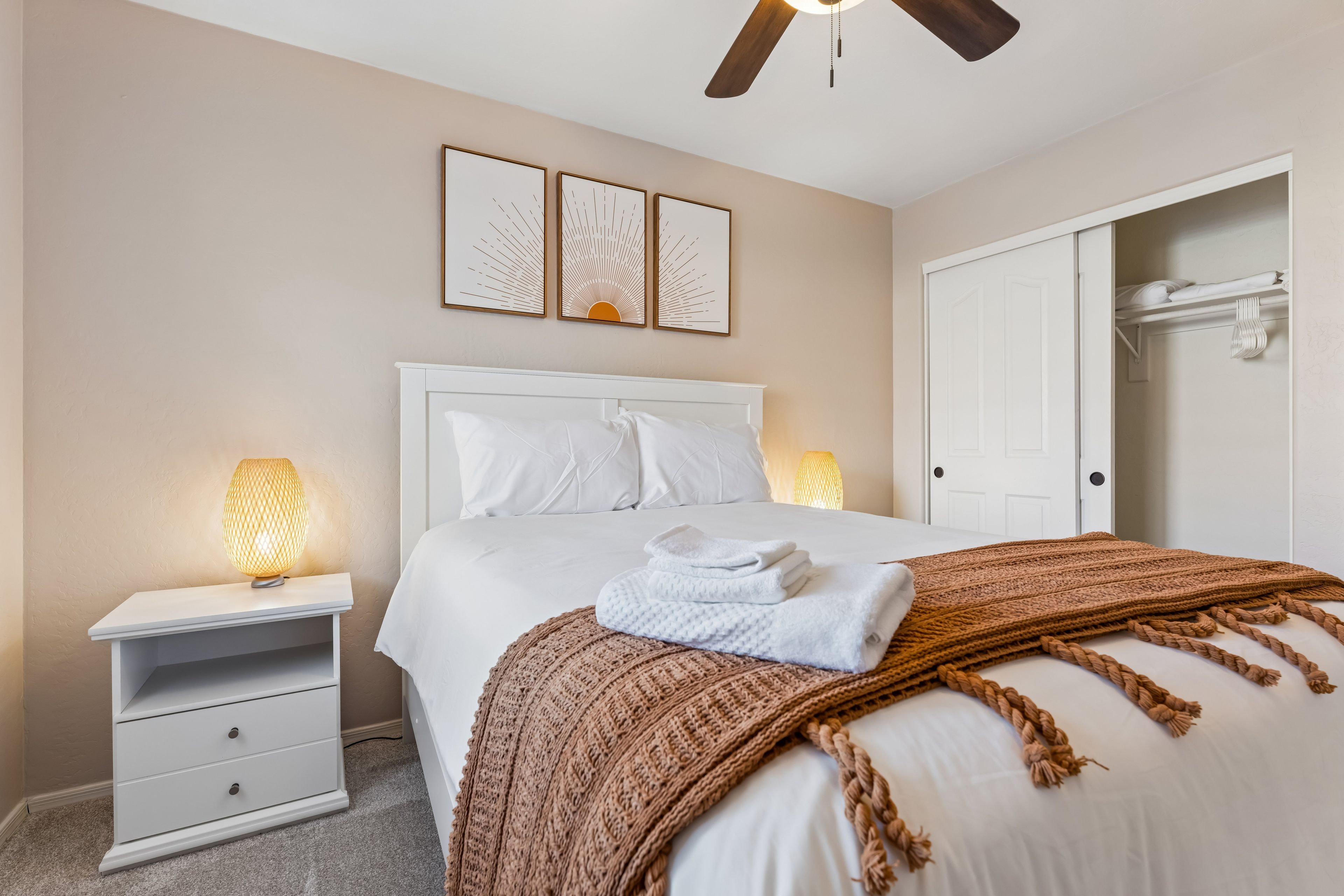 A neatly made bed with a white comforter and pillows, a brown knitted throw, a bedside lamp, and a white nightstand with drawers.