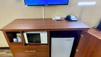 A hotel room with a television displaying a schedule, a desk with a telephone, and a built-in mini-fridge.