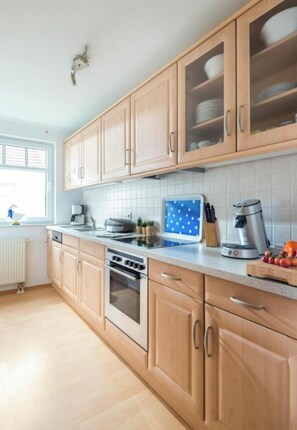 A kitchen with wooden cabinets, a built-in oven, a coffee maker, and a window with blinds. - Marienstrasse 4a Apartment 4 (Norderney)