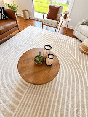 A living room with a round wooden coffee table, a white rug, a brown leather sofa, and a view of a green lawn through a glass door.