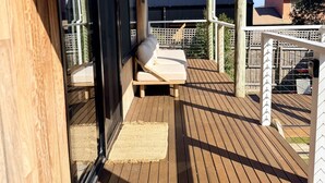 Una terrassa de fusta amb una porta corredissa de vidre que dona a un balcó, una cadira còmoda amb coixins i una catifa petita.