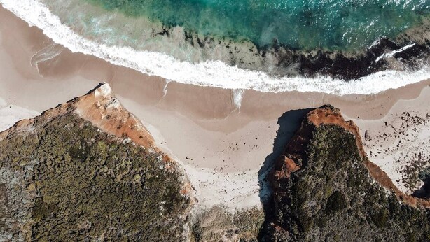 Vista aèria d'una platja amb aigües turqueses cristal·lines, sorra daurada i roques.