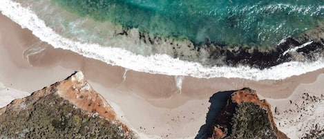 An aerial view of a beach with clear turquoise waters, sandy shore, and rocky outcrops.