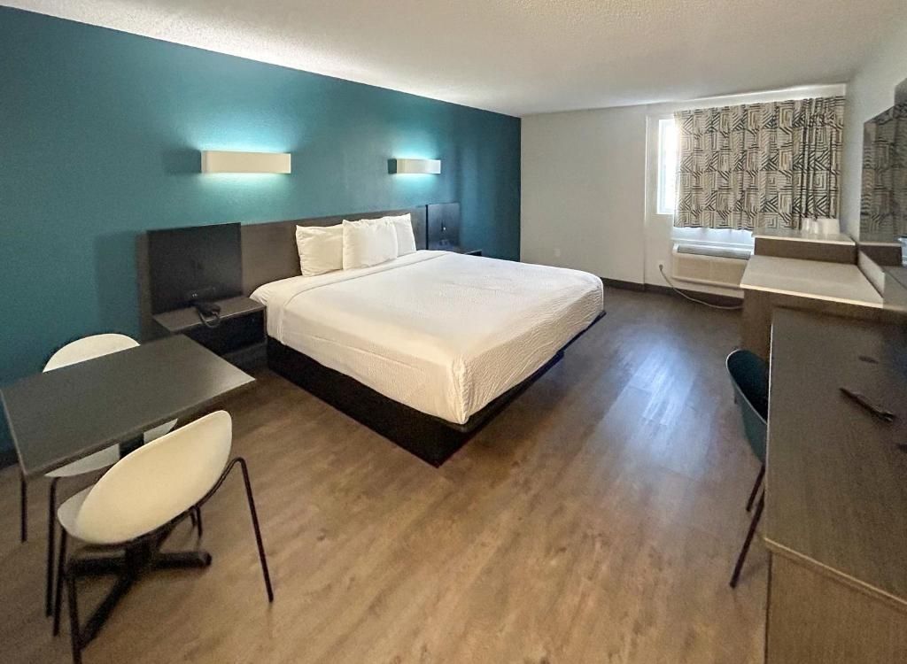 A hotel room with a bed, a desk, a chair, a nightstand, and a window with patterned curtains.