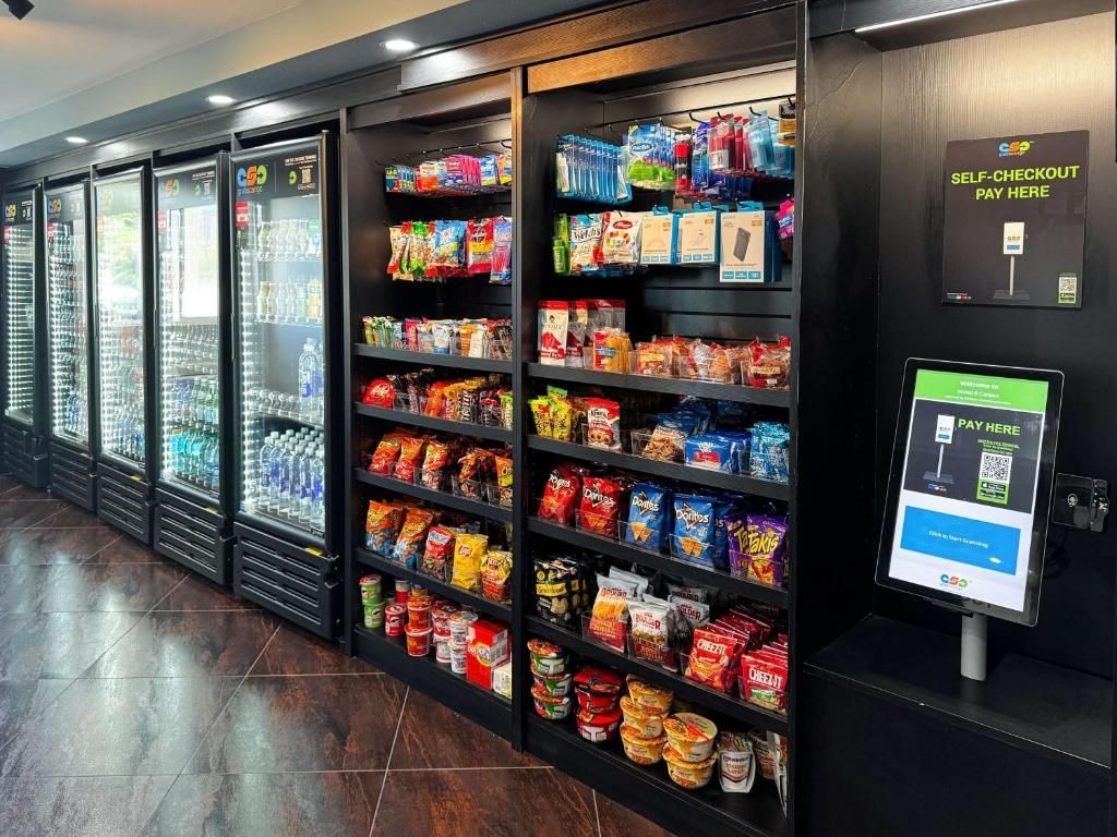 Un área de autoservicio con un terminal de pago digital y estantes llenos de aperitivos y bebidas.