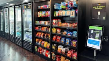 Un área de autoservicio con un terminal de pago digital y estantes llenos de aperitivos y bebidas.