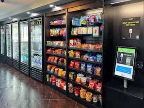 Un área de autoservicio con un terminal de pago digital y estantes llenos de aperitivos y bebidas.