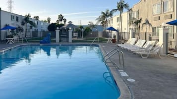 A swimming pool with lounge chairs and umbrellas, surrounded by a fence and palm trees.