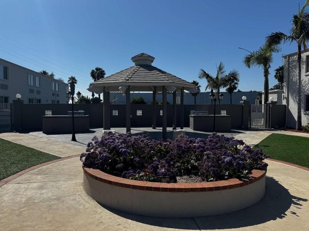 A gazebo with a tiled roof, surrounded by a circular flower bed with purple flowers, set against a backdrop of a gated area with palm trees and a building.