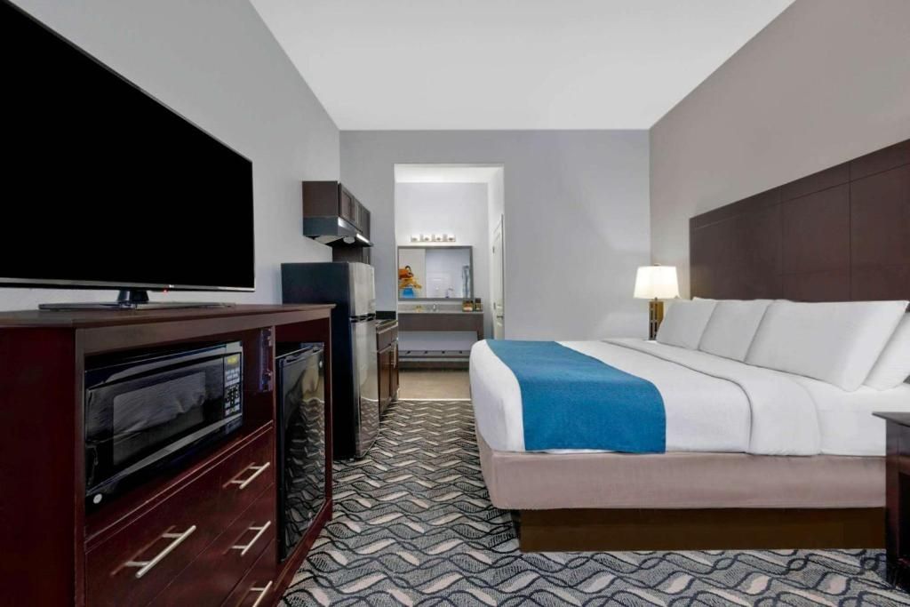 Una habitación de hotel con una cama, televisión, escritorio con lámpara y kitchenette.