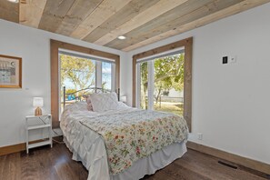 A bedroom with a bed, a bedside table, a lamp, and large windows. - Peaceful 3-bedroom lake house with hot tub, and sauna.Birders paradise. (Amherstburg)
