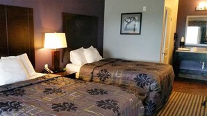 A hotel room with two beds, a bedside table with a lamp, a mirror, and a bathroom with a sink and mirror. - Your Budget-Friendly Home Away from Home l 2 Queen Beds Suite (Beeville)
