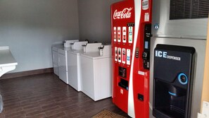 A room with a Coca-Cola vending machine, an ice dispenser, and a white appliance, possibly a coffee maker or tea maker.