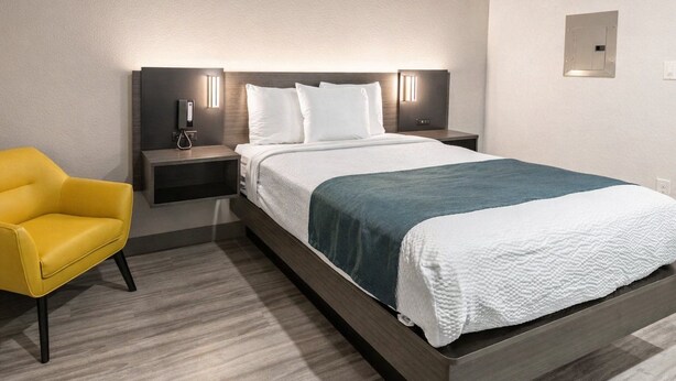 Una camera d'hotel con un letto, comodini, una lampada, una sedia e una presa elettrica.