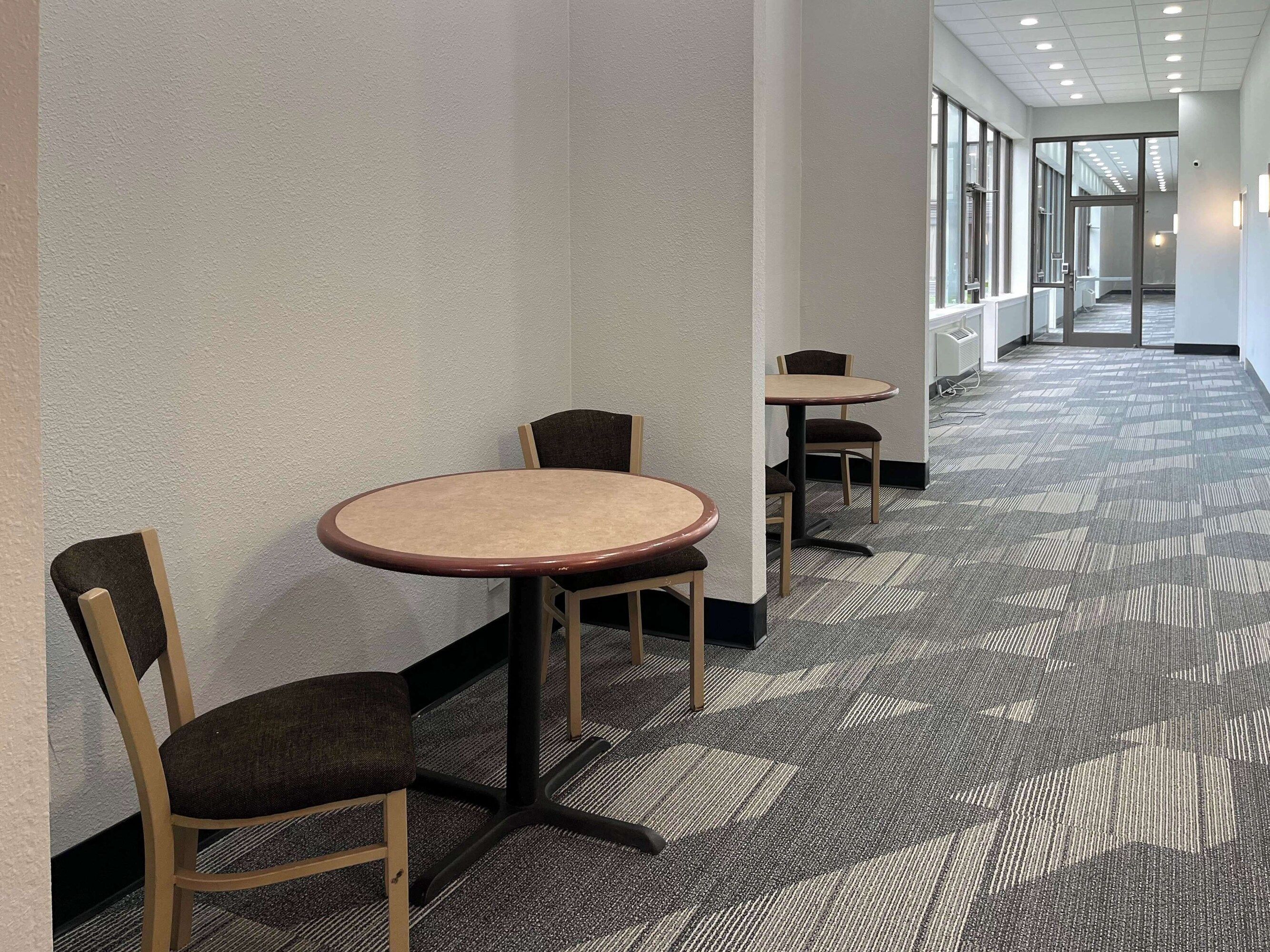 Un couloir doté d’un revêtement de sol en moquette, une série de tables et de chaises, et une porte vitrée donnant sur un autre espace.