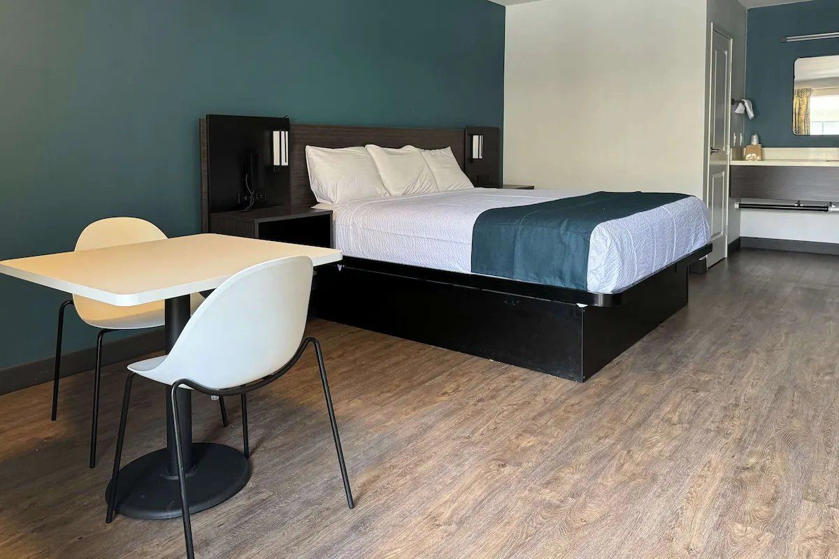 A modern hotel room with a bed, bedside table, chair, and a small kitchenette area.