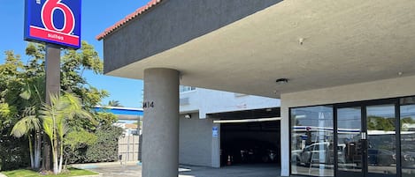 L’entrée d’un hôtel, avec une enseigne indiquant « 6 suites », un espace de stationnement couvert et un bâtiment à la façade en béton.