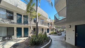 Une cour intérieure avec un jardinière centrale, un palmier et un bâtiment doté de balcons et de portes numérotées 220 et 203.