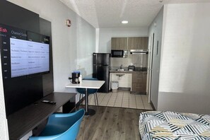 Camera d'albergo compatta con televisore che mostra una guida ai canali, un piccolo tavolo con sedie, angolo cottura e un letto.